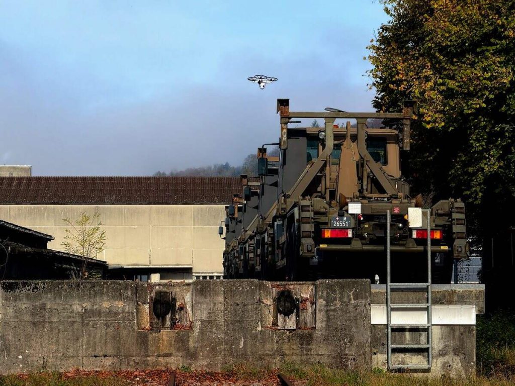 Dron en vuelo de vigilancia sobre vehículos industriales y muro perimetral de instalación.